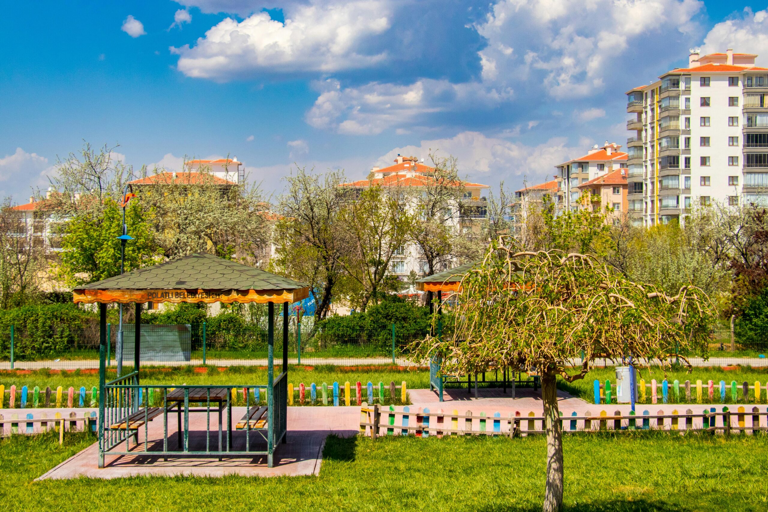 Bright urban park scene with gazebo, green trees, and modern buildings under a blue sky.